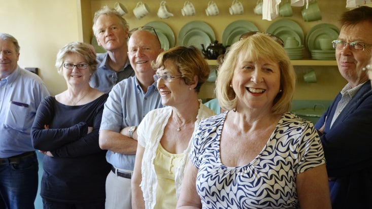 A group of people looking interestedly to the left of the camera at something indoors with shelves of plates behind them